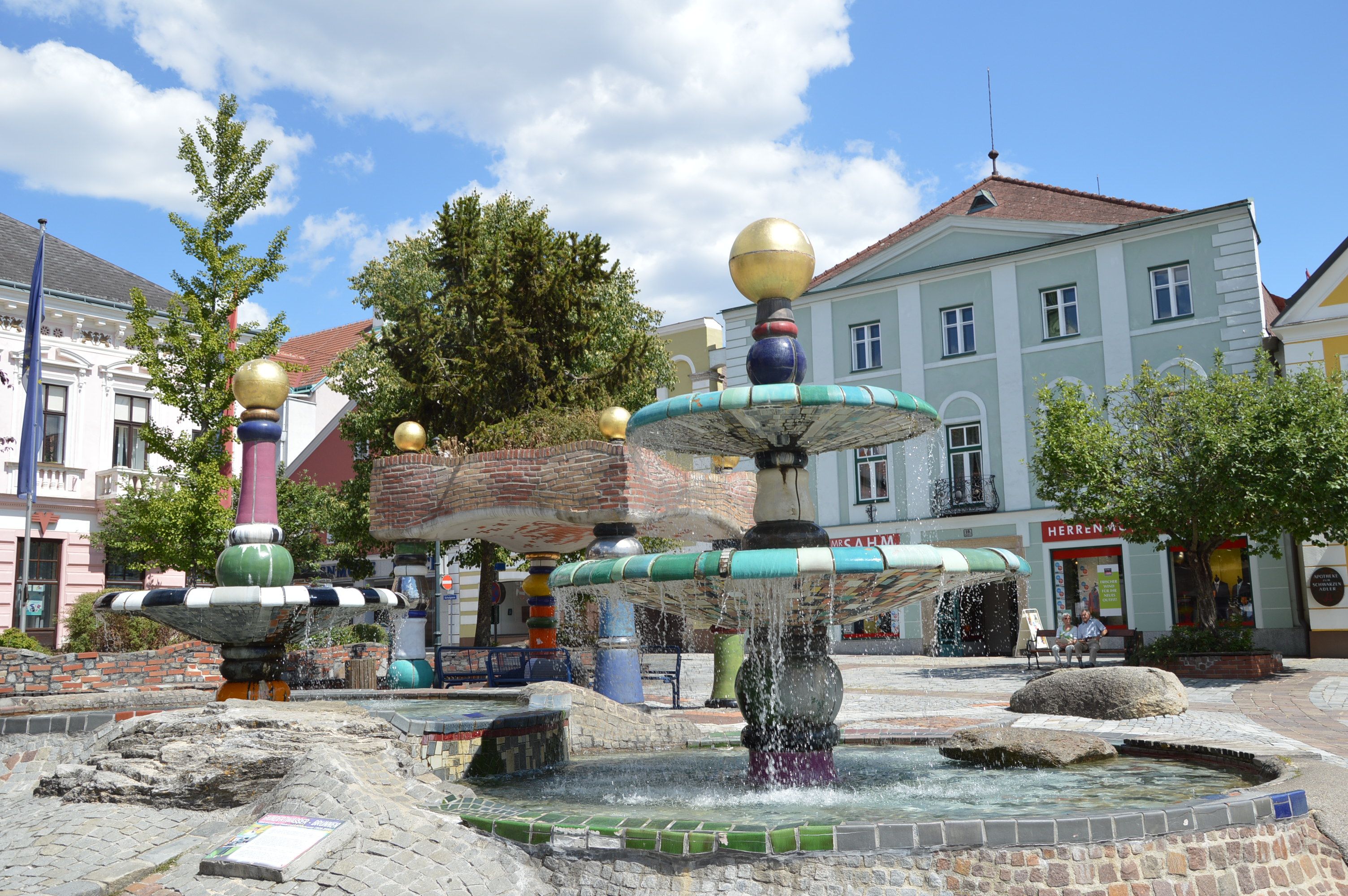 Színes Hundertwasser-stílusú szökőkút aranyszínű gömbökkel és folyóvízzel, épületekkel és fákkal körülvéve.