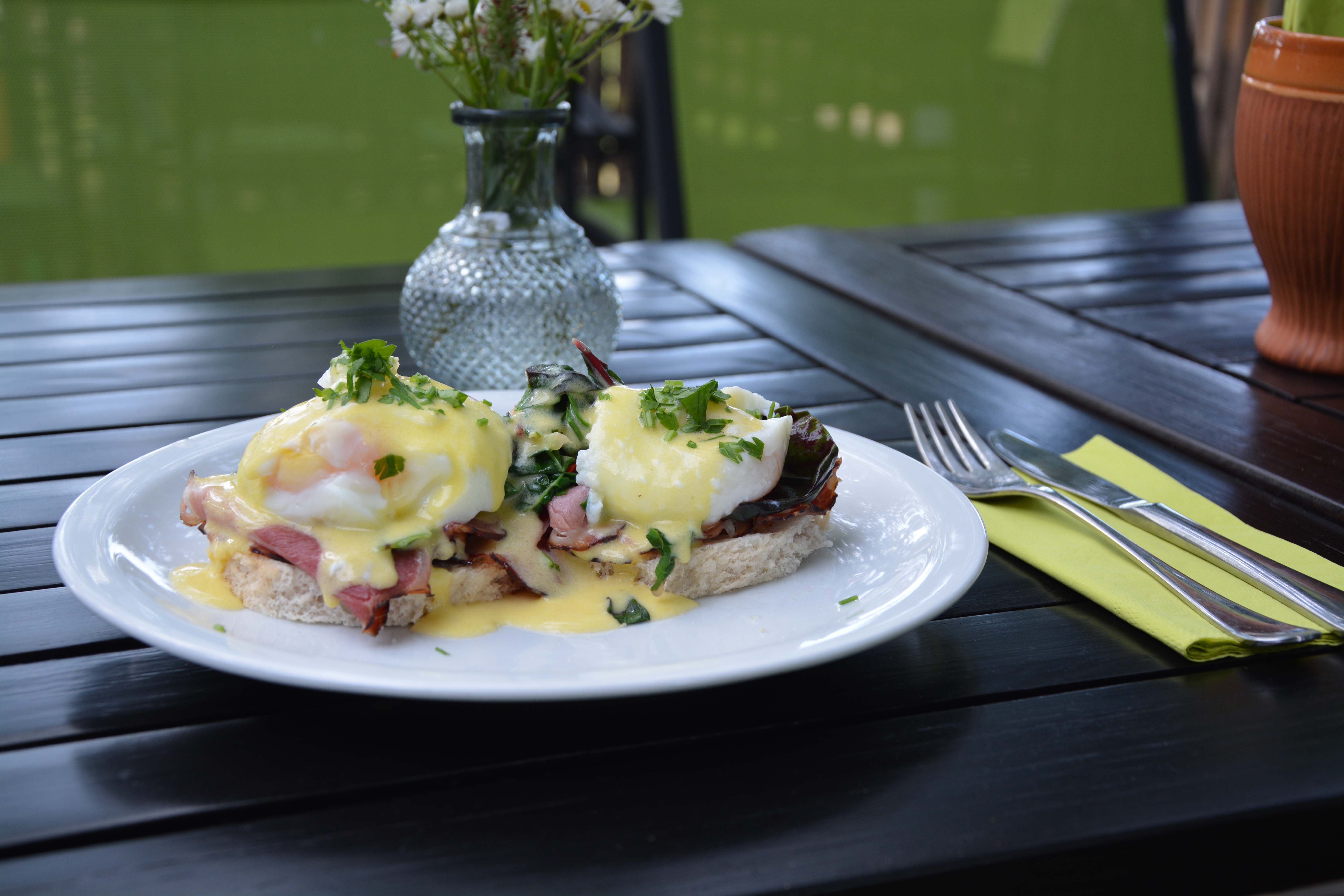 Egy tányér tojás Benedict egy faasztalon, mellette evőeszközökkel és egy virágvázával.