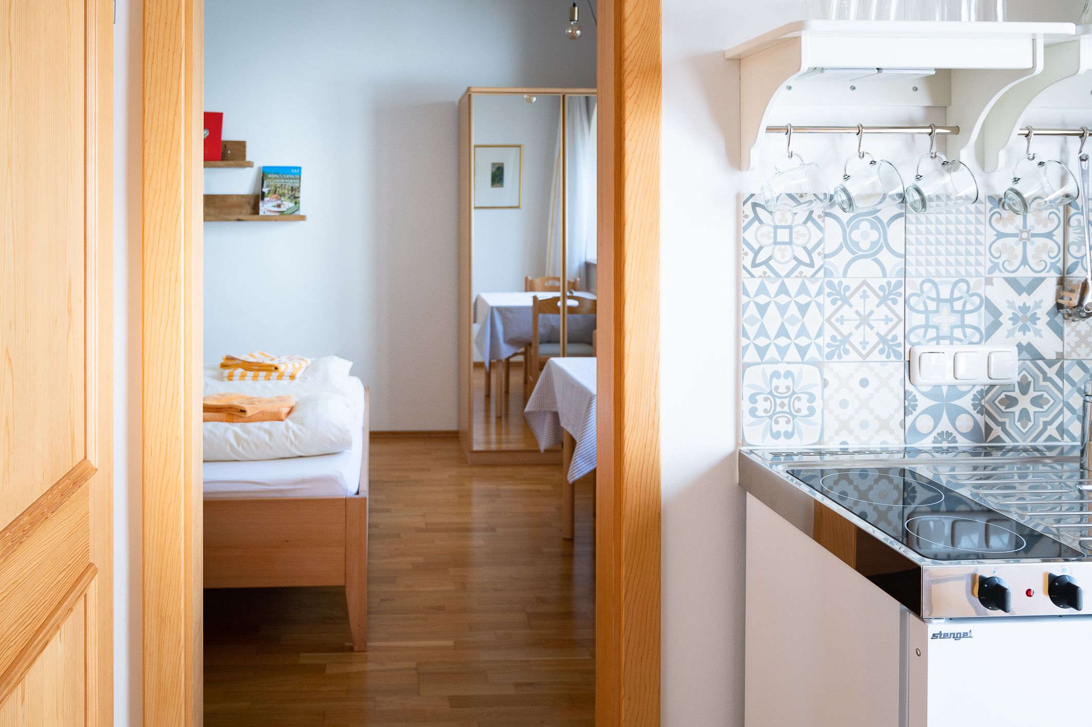 Egy szoba belső nézete, ágy, tükör, asztal és egy kis konyha mintás csempével.