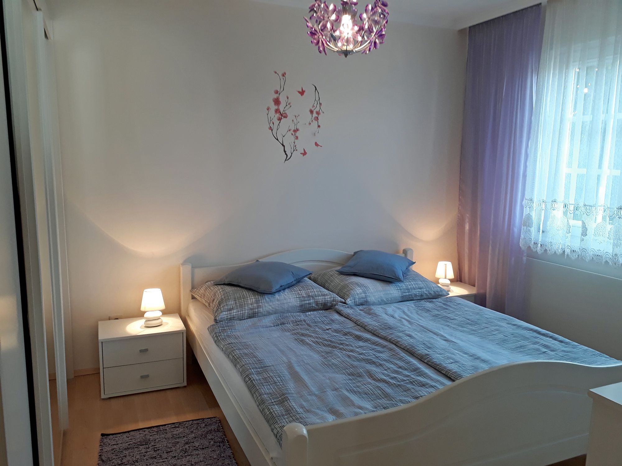 Hangulatos hálószoba franciaággyal, két éjjeliszekrénnyel, lámpákkal és lila függönyökkel.