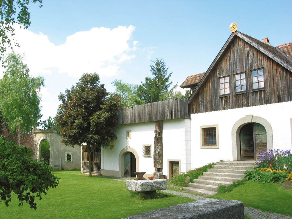 Hagyományos épület, fából készült tetővel és fehér homlokzattal, zöld kerttel körülvéve.