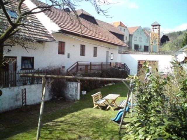 Vidéki udvar fehér épületekkel, fa asztal és székek a kertben, hinta és csúszda az előtérben.