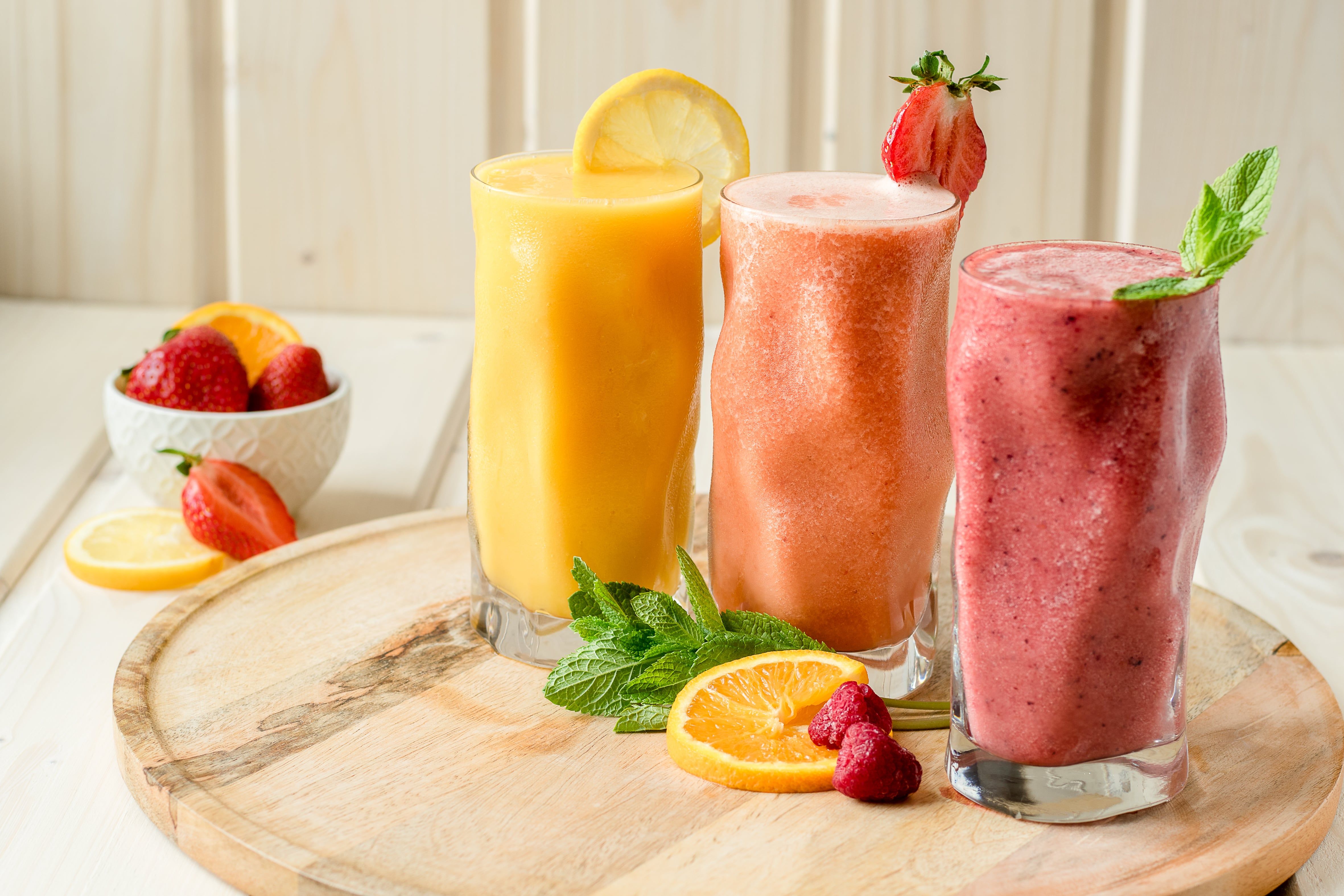 Három smoothie pohárban, fából készült deszkán, gyümölcsökkel és mentával díszítve.