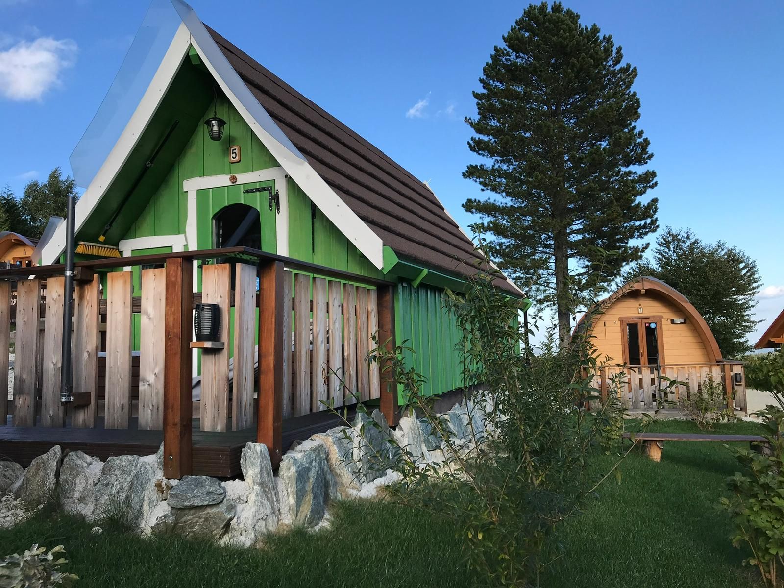 Két kis faházikó glamping stílusban, zöld és barna homlokzattal, fákkal és fűvel körülvéve.