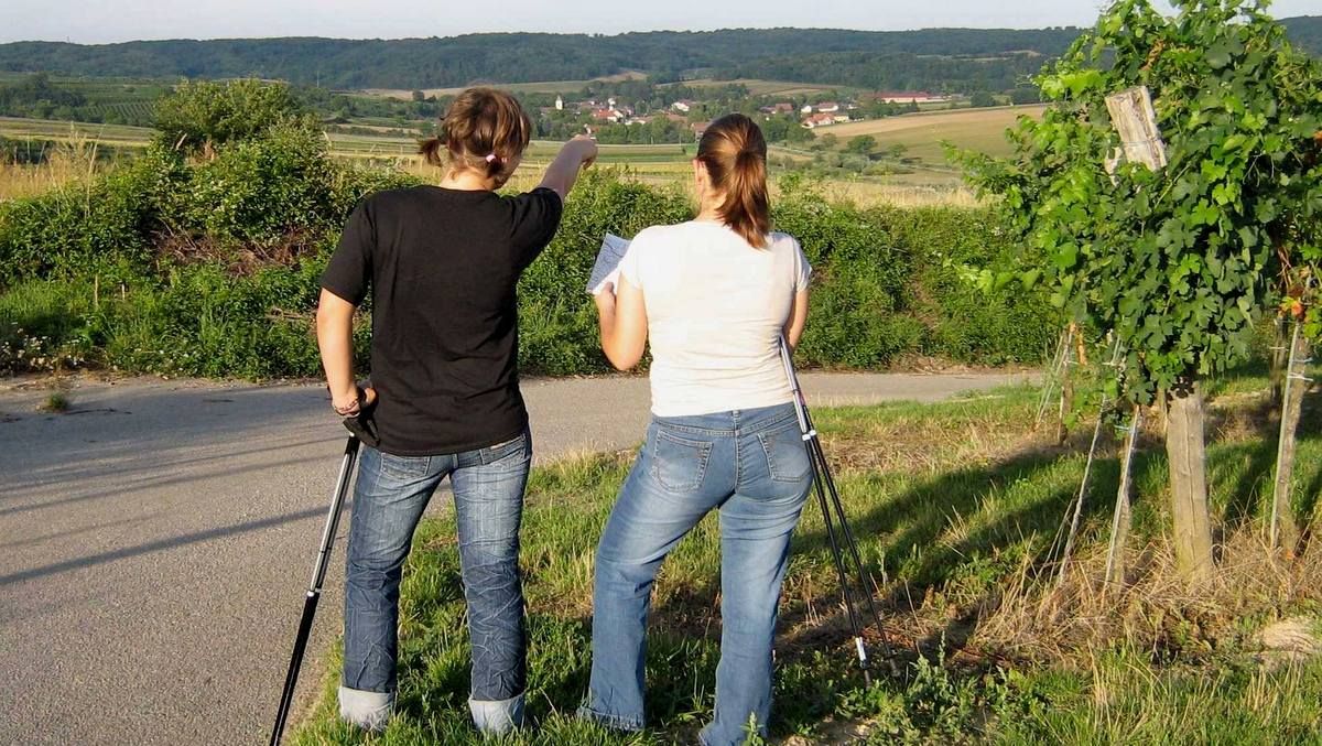 Két sétapálcás nő áll egy ösvényen, és a távolba néznek, az egyikük egy falu irányába mutat.
