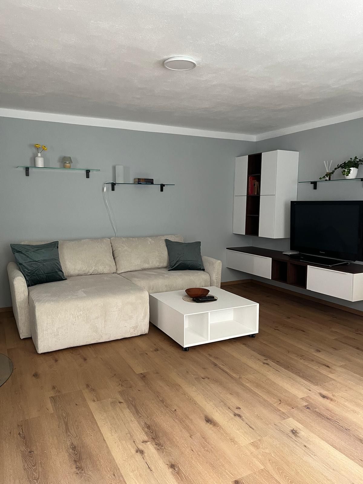 Modern nappali bézs színű kanapéval, fehér dohányzóasztallal, fapadlóval és fali polcokkal.