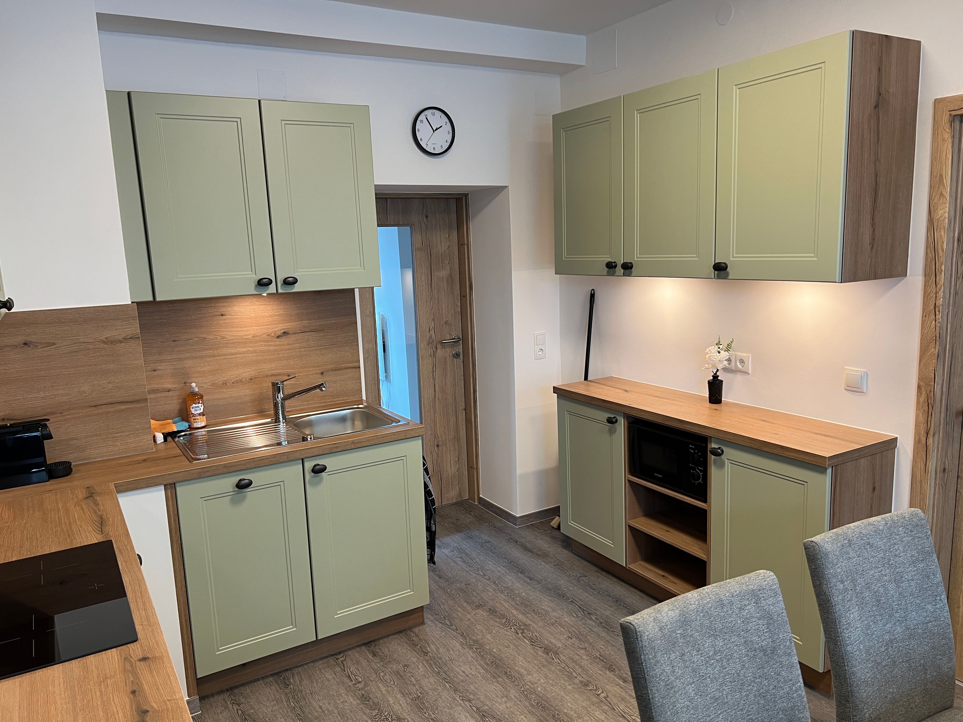 Modern konyha zöld szekrényekkel, fa munkalapokkal és szürke székekkel.