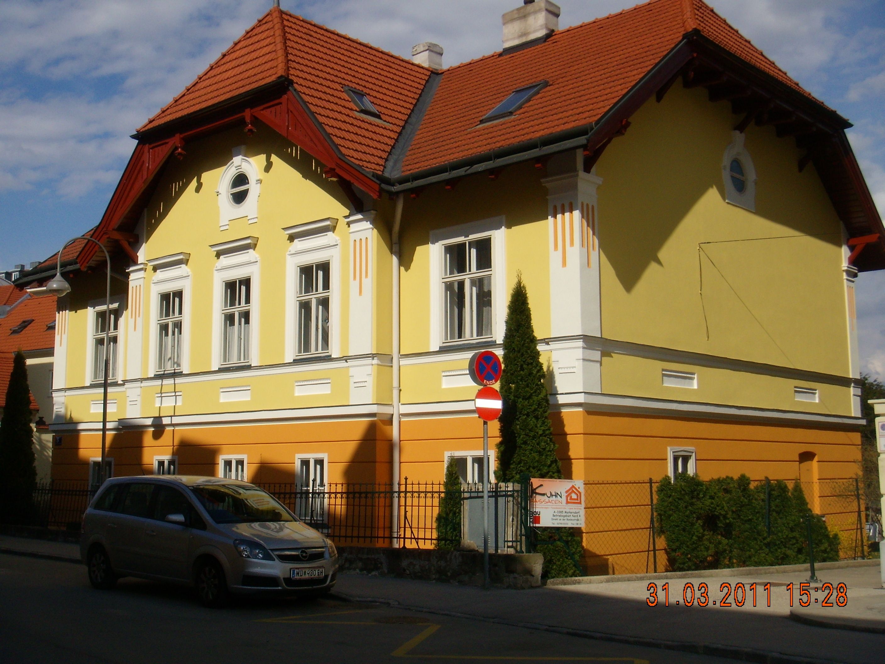 Sárga és narancssárga épület piros tetővel és egy parkoló autóval előtte.