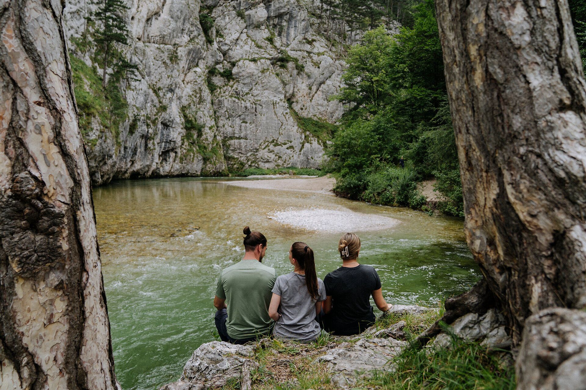 Drei Personen sitzen am Ufer der Scharzach im Höllental am Wanderweg 1. Wiener Wasserleitungsweg und machen Pause. Rundum sie ragen Felden und Bäume in die Höhe. 