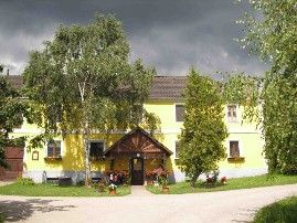 Sárga, fából készült tetővel és kerttel rendelkező, fákkal körülvett ház.