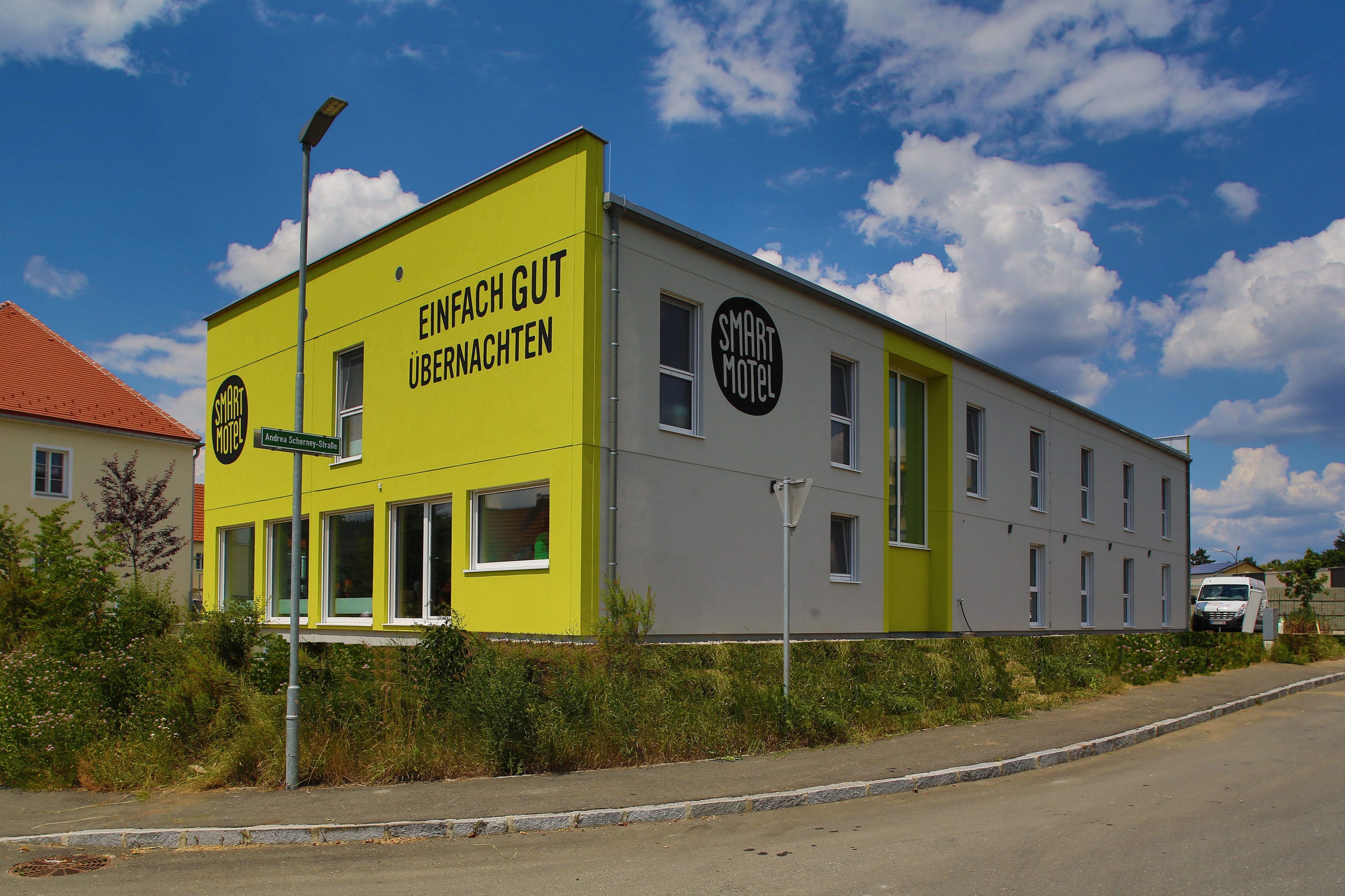 Egy modern, kétszintes motel külső nézete, sárga homlokzattal és az "Einfach gut übernachten" felirattal.