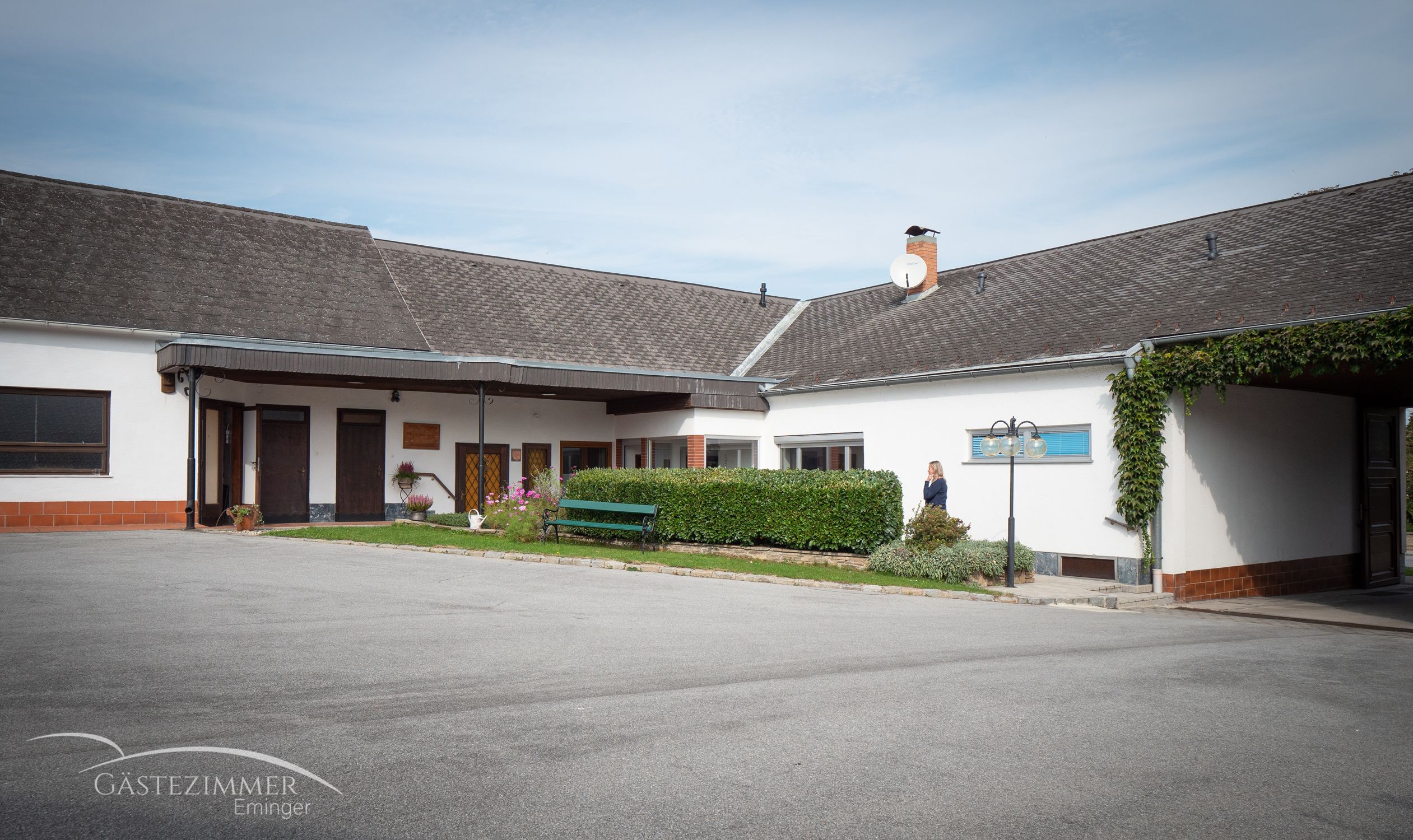 Egy udvar egy épülettel, parkolóhelyekkel és egy sövénnyel. Egy személy áll a sövény mellett. Az épületen egy parabolaantenna látható.