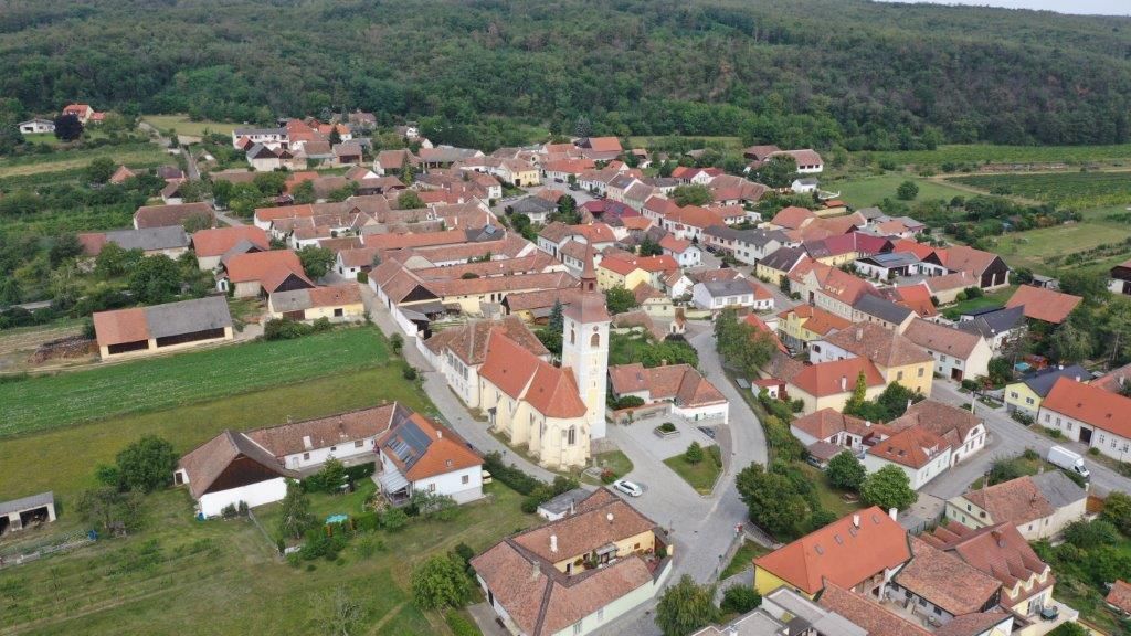 Légi felvétel egy kis faluról, amelynek központi temploma vörös tetőkkel van körülvéve, zöld mezőkkel és erdőkkel.