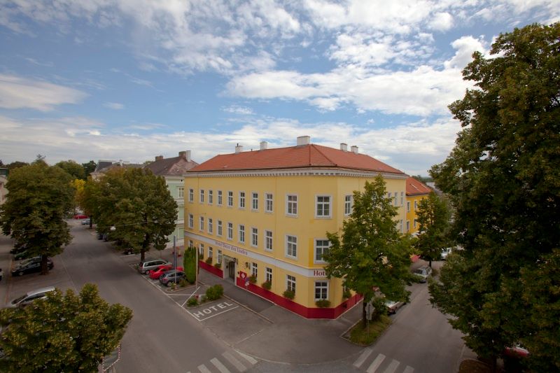Külső nézet egy sárga színű, piros tetős hotelről, amelyet fák és utcák vesznek körül.