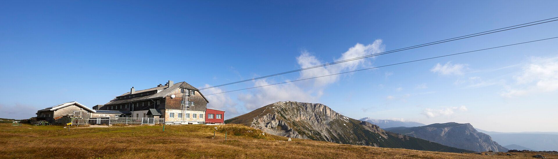 Panorámakép egy hegyi kunyhóról a környező hegycsúcsokkal a kék ég alatt.