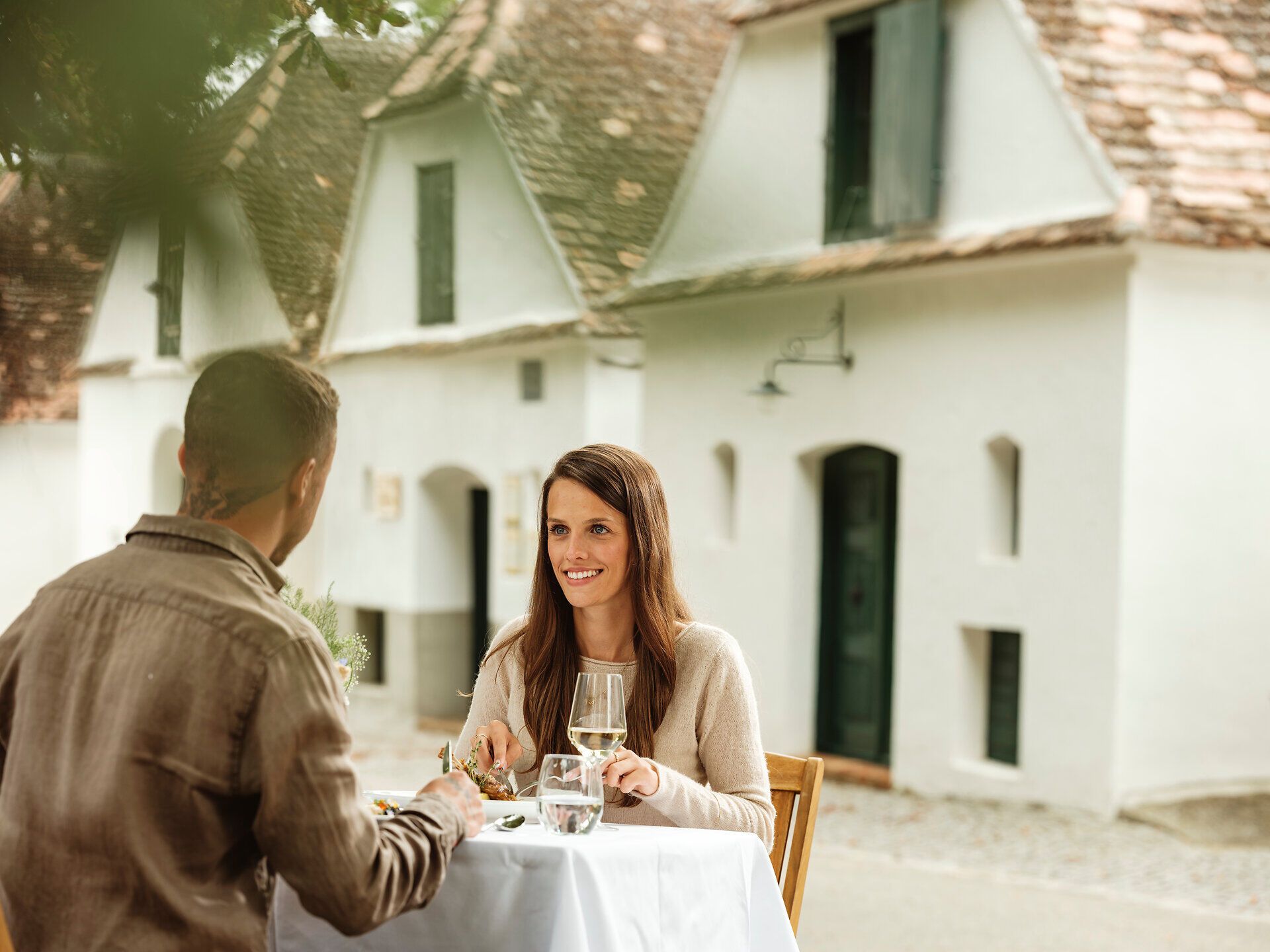 In der malerischen Kellergasse Zipf genießen Paare ein romantisches Dinner unter freiem Himmel, umgeben von den sanften Hügeln des Weinviertels. Die Atmosphäre ist geprägt von der warmen Abendsonne, die die charmanten Weinhäuser in goldenes Licht taucht und die Vorfreude auf kulinarische Köstlichkeiten weckt.