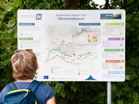 Egy szem&eacute;ly n&eacute;zi az als&oacute;-ausztriai Tulln r&eacute;gi&oacute;ban tal&aacute;lhat&oacute; kerti ker&eacute;kp&aacute;rt&uacute;r&aacute;kr&oacute;l sz&oacute;l&oacute; inform&aacute;ci&oacute;s t&aacute;bl&aacute;t, amelyen t&eacute;rk&eacute;p &eacute;s k&uuml;l&ouml;nb&ouml;ző &uacute;tvonalakr&oacute;l sz&oacute;l&oacute; inform&aacute;ci&oacute;k l&aacute;that&oacute;k.