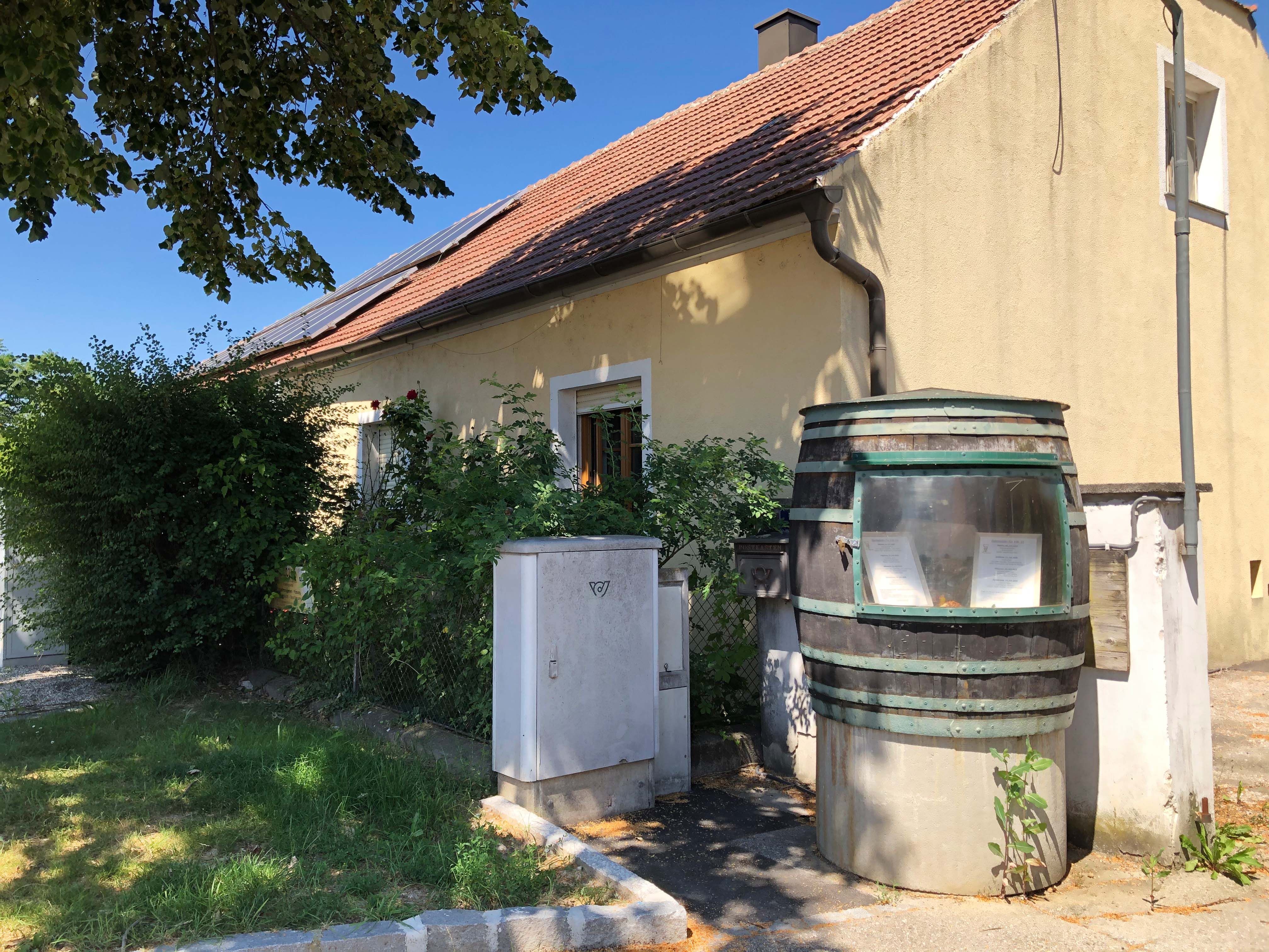 Sárga ház piros cseréptetővel és napelemekkel. Az előtérben egy nagy hordó látóüveggel és növényekkel.