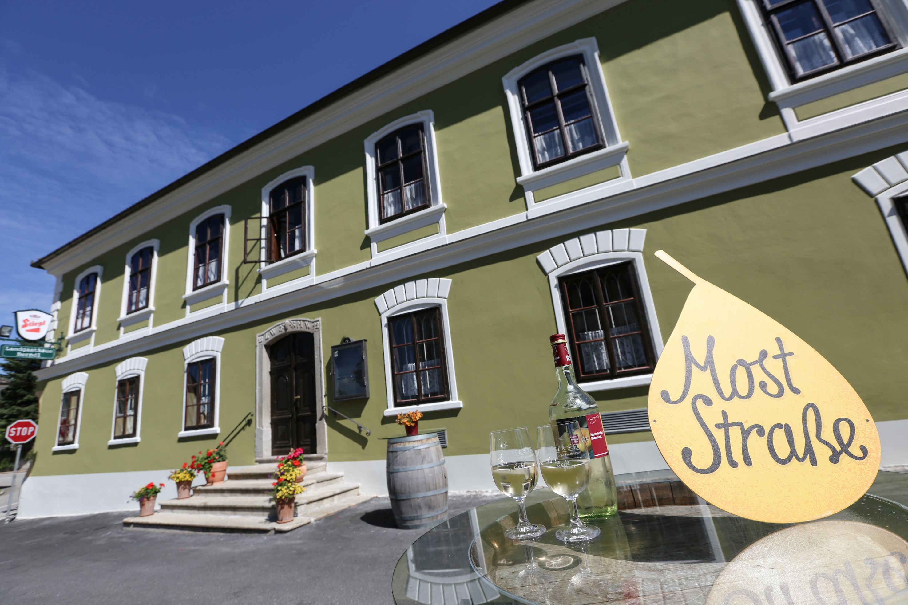 Zöld épület fehér ablakokkal, előtte asztal egy üveg gyümölcsborral és poharakkal, felirat: "Gyümölcsbor Straße".