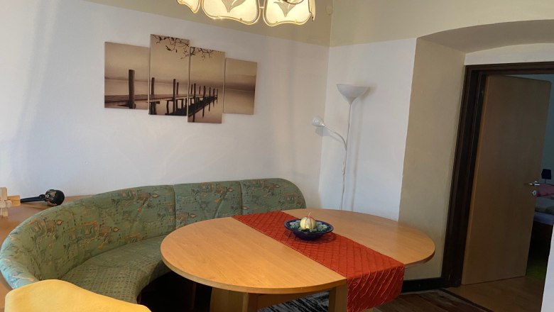 Egy nappali egy ov&aacute;lis faasztallal, egy z&ouml;ld sarokpaddal, egy piros asztali fut&oacute;val &eacute;s egy k&eacute;ppel a falon. A sarokban &aacute;ll egy &aacute;ll&oacute;l&aacute;mpa.