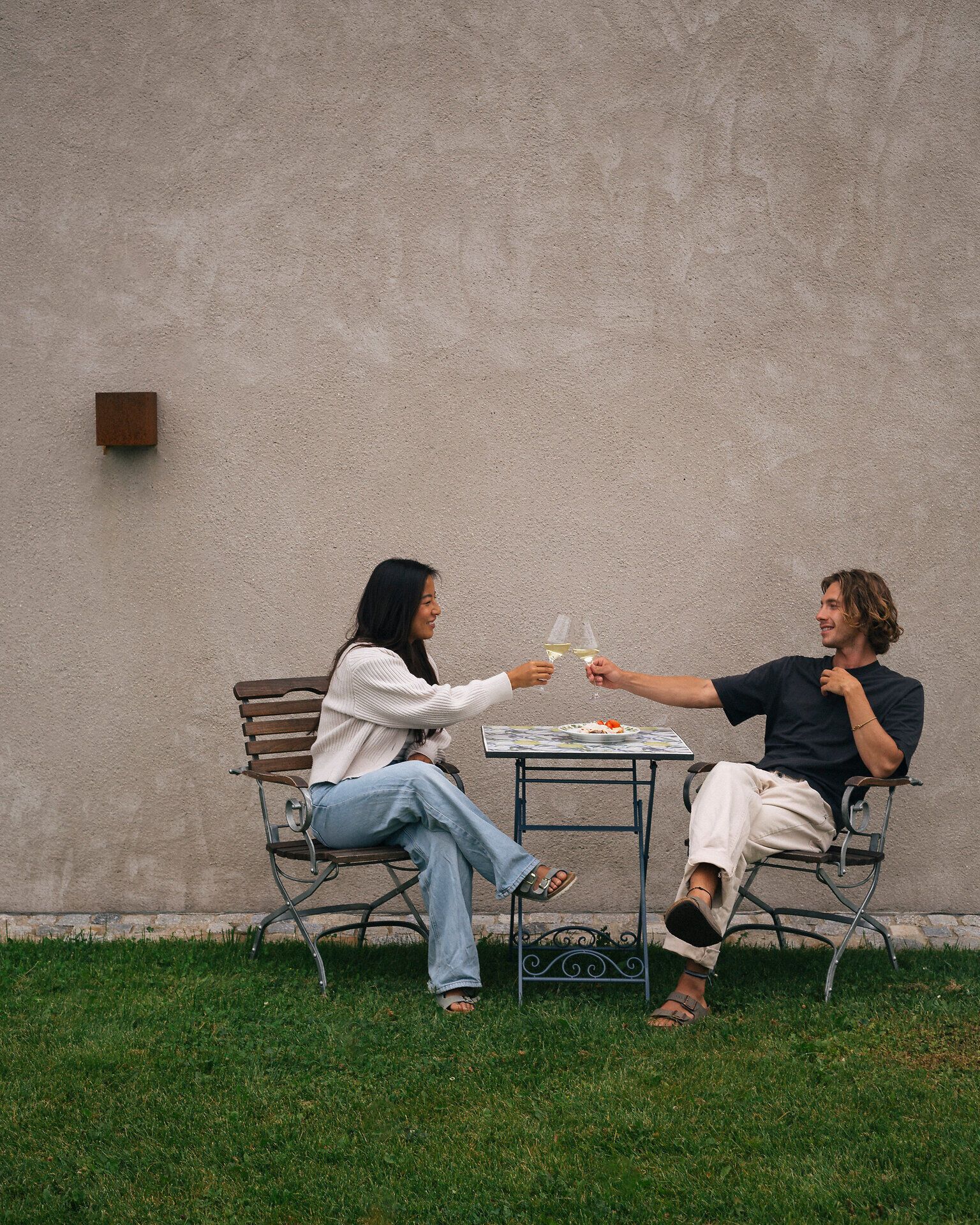 Zwei Personen sitzen im Garten an einem kleinen Tisch vor einer hellen Wand und stoßen mit Weißweingläsern an.