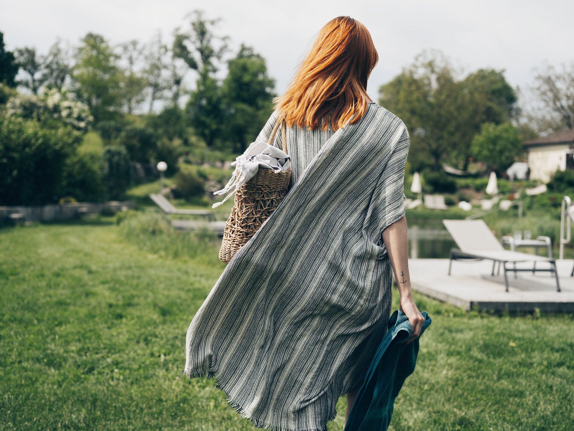 Eine Frau mit rotem Haar schlendert entspannt durch die grüne Wiese, während sie einen geflochtenen Korb trägt. Die sanften Hügel im Hintergrund und die blühenden Bäume schaffen eine idyllische Atmosphäre, die zum Verweilen einlädt.