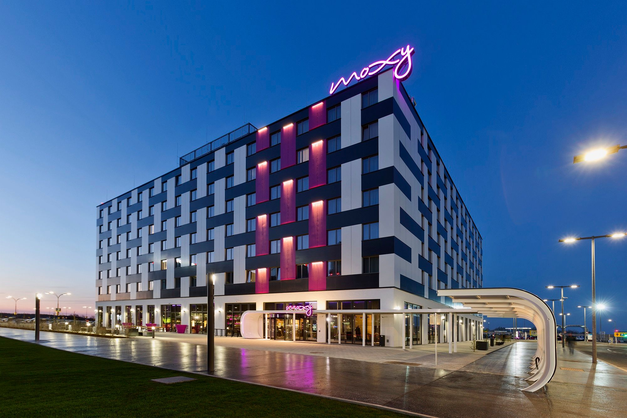 A Moxy Vienna Airport Hotel külső nézete alkonyatkor, kivilágított feliratokkal.