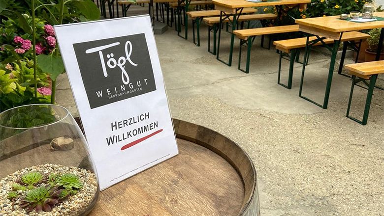 Egy boroshordón a "Weingut Tögl" és a "Herzlich Willkommen" ("Üdvözöljük") feliratú tábla áll. A háttérben sörasztalok és növények láthatók.