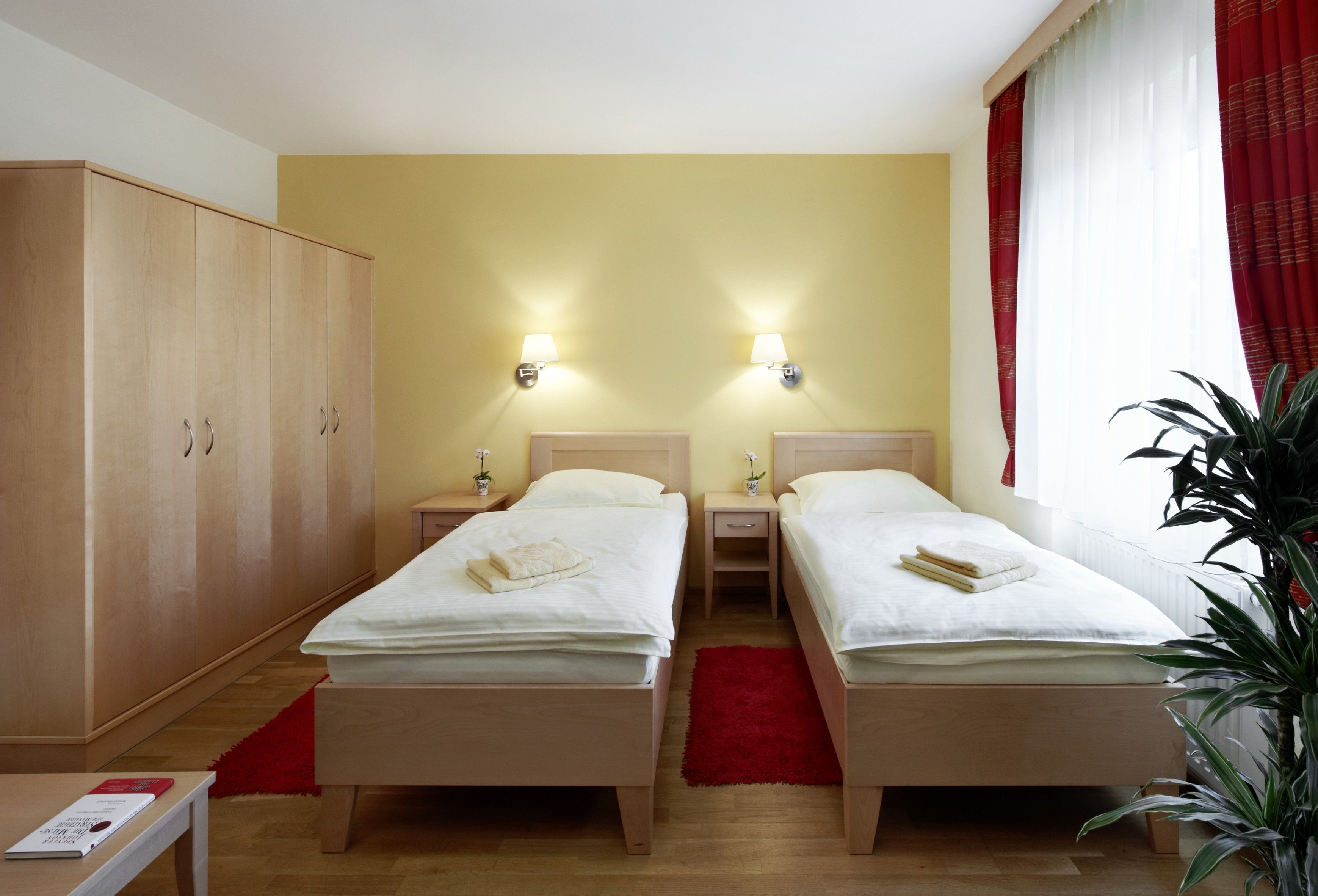 Vendégszoba két egyszemélyes ággyal, nagy szekrénnyel, piros függönyökkel és egy növénnyel.