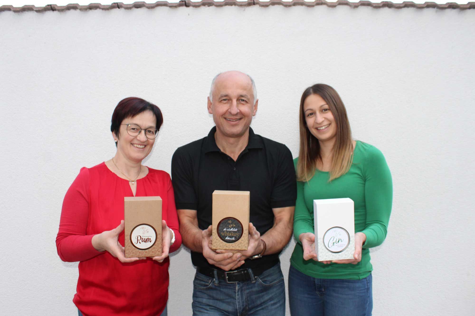 Három ember rumot, whiskyt és gint tartalmazó ajándékdobozokat tart egy fehér fal előtt.