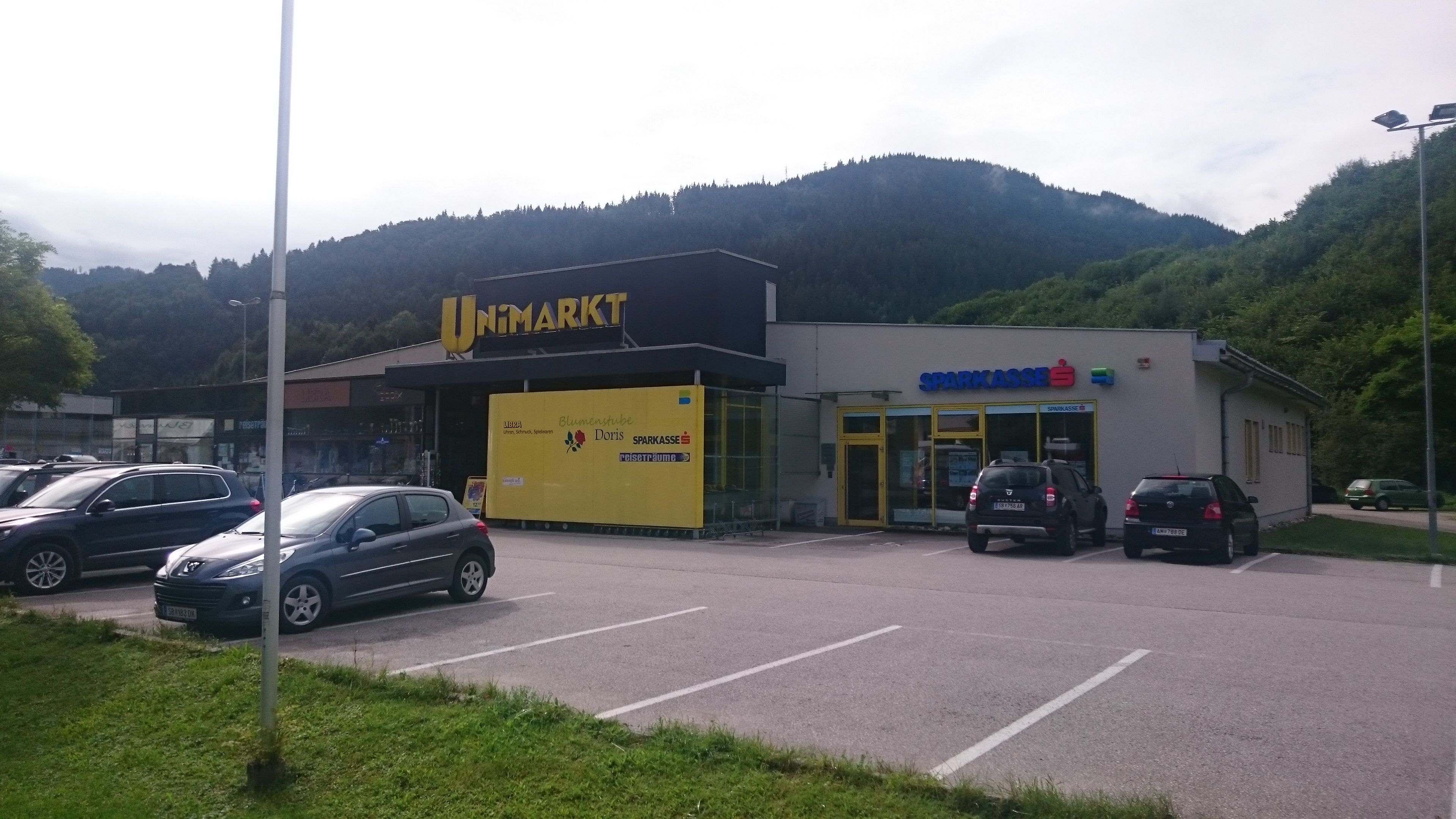 Bevásárlóközpont Unimarkt szupermarkettel és takarékpénztárral az erdős dombok előtt.