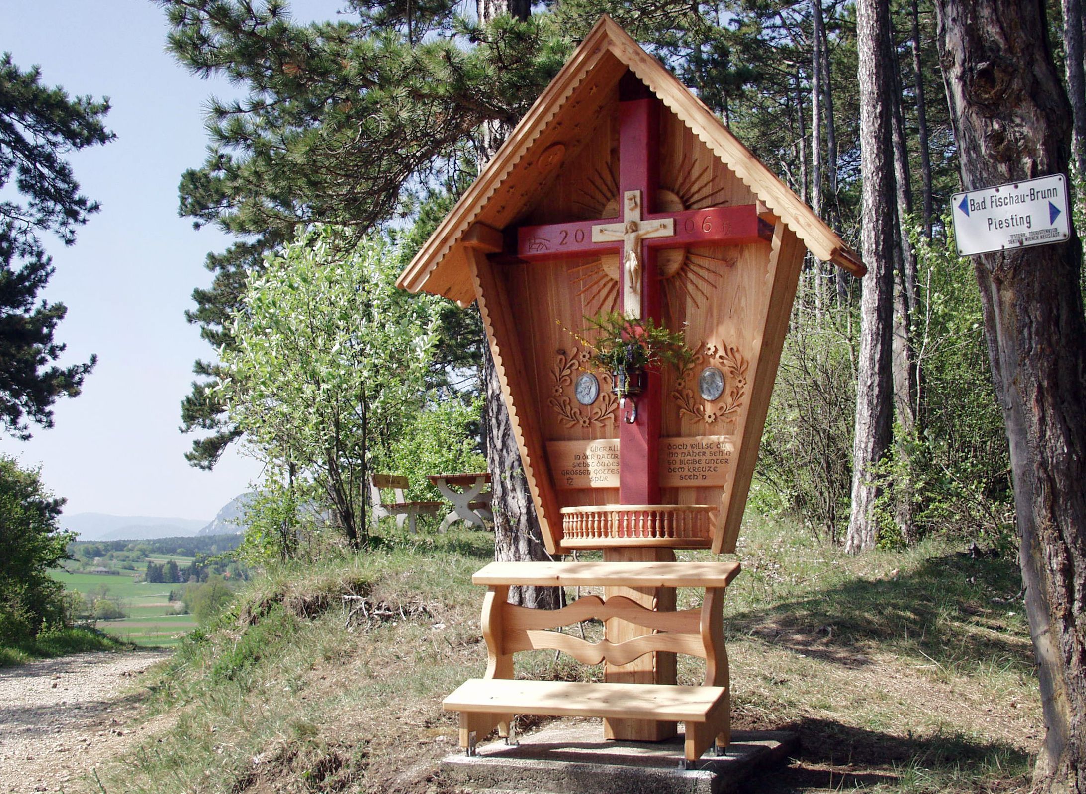Fakereszt vörös gerendával és faragott tetővel erdei tájban.
