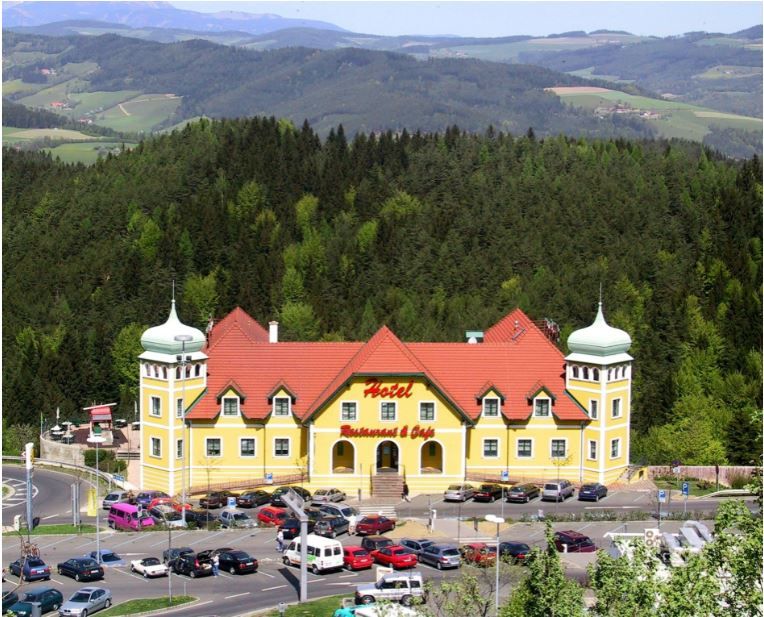 Sárga szálloda piros tetőkkel zöld, dombos tájban.