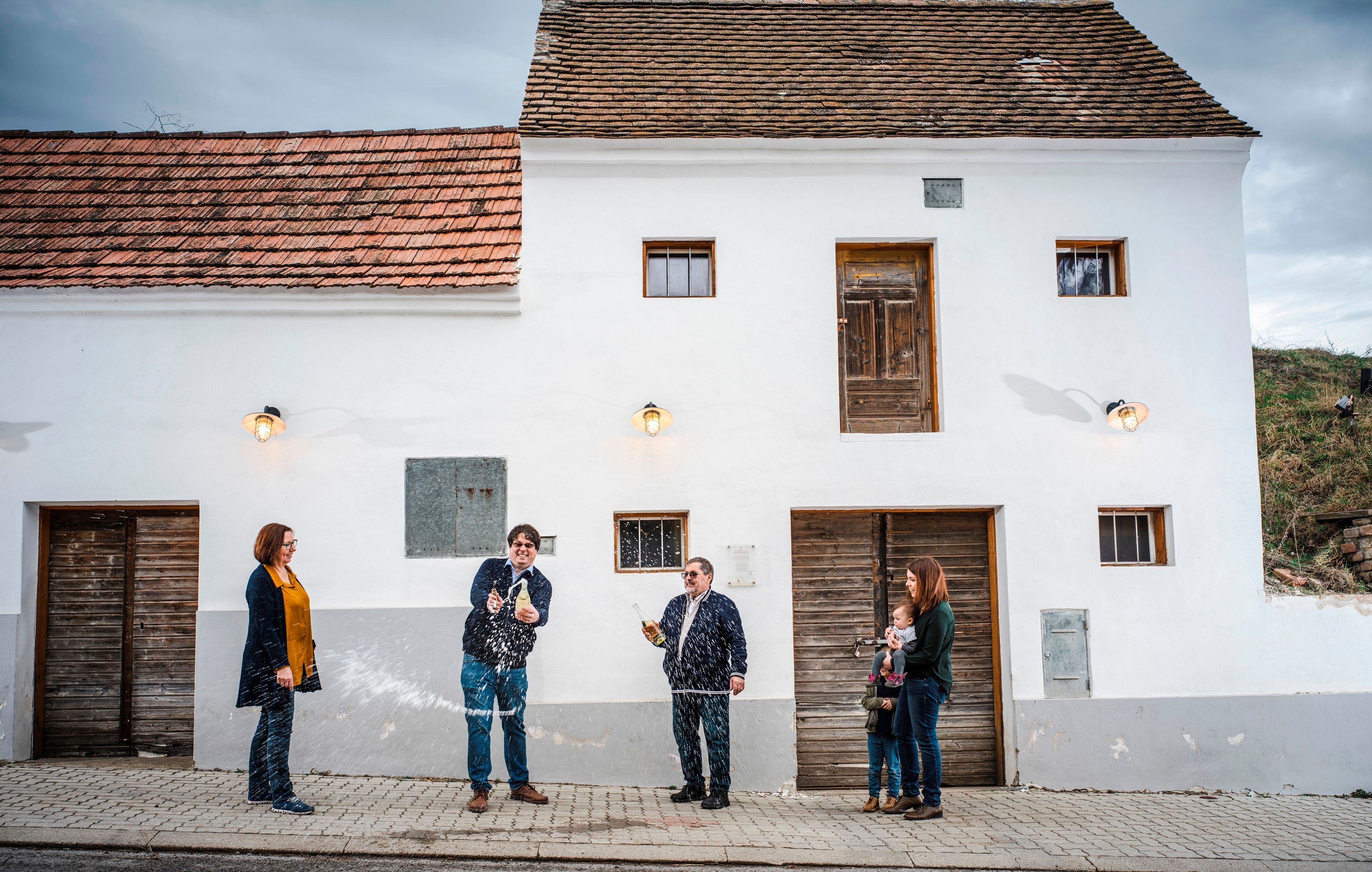 Emberek egy csoportja áll egy fehér épület előtt, miközben az egyik személy egy palackot permetez.