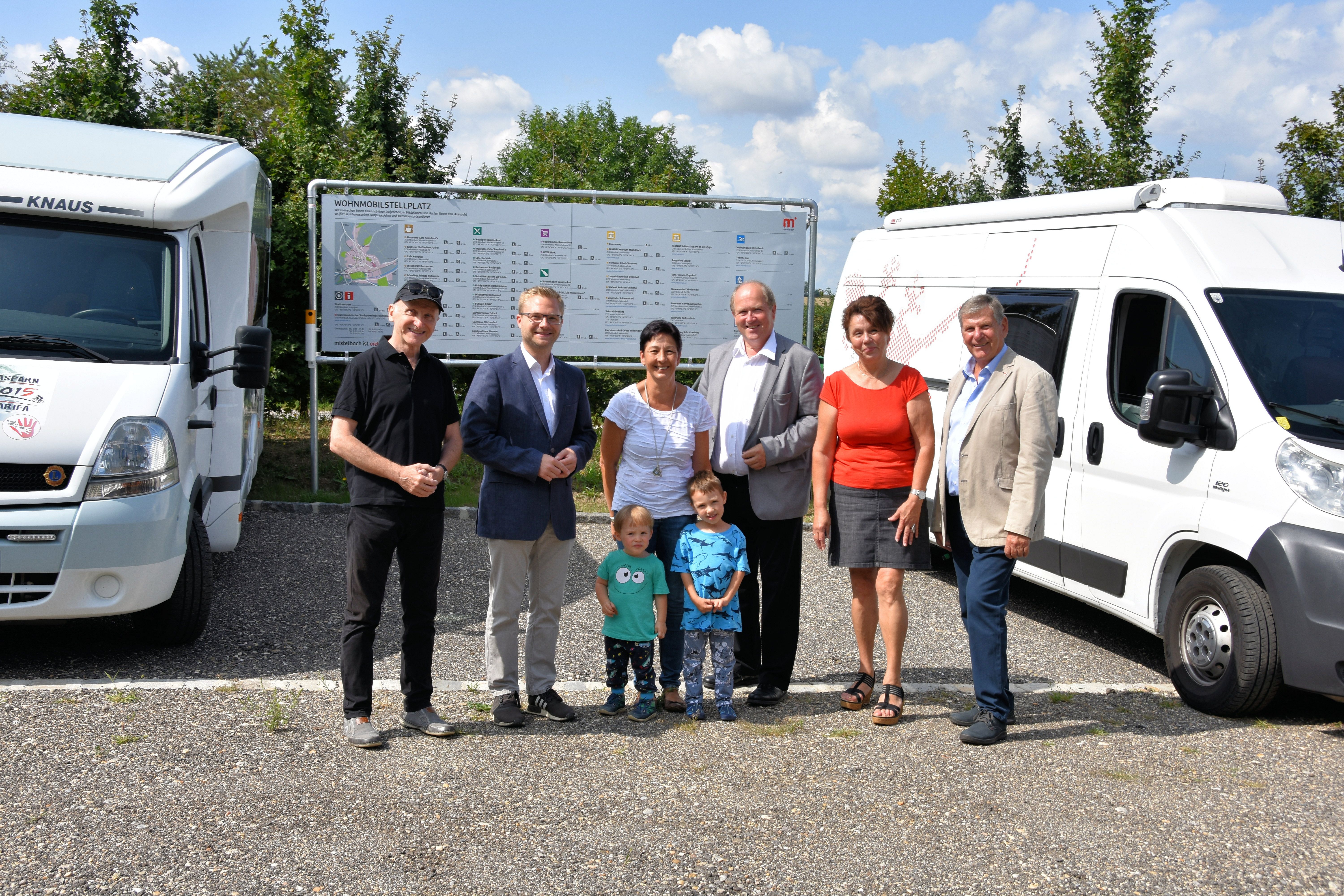 Emberek csoportja a lakóautók előtt és egy tábla egy pályán.