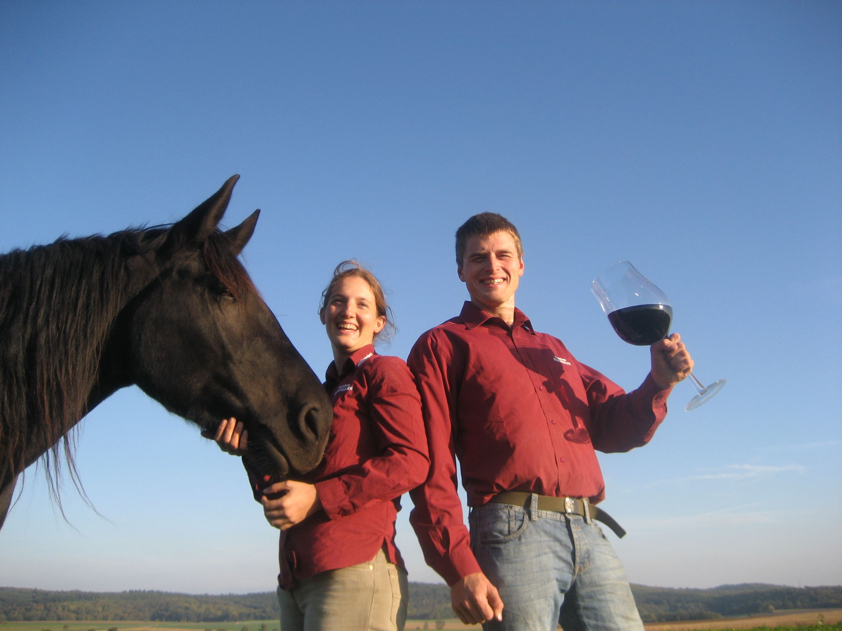 Két piros inges ember egy fekete lóval és egy nagy borospohárral odakint.