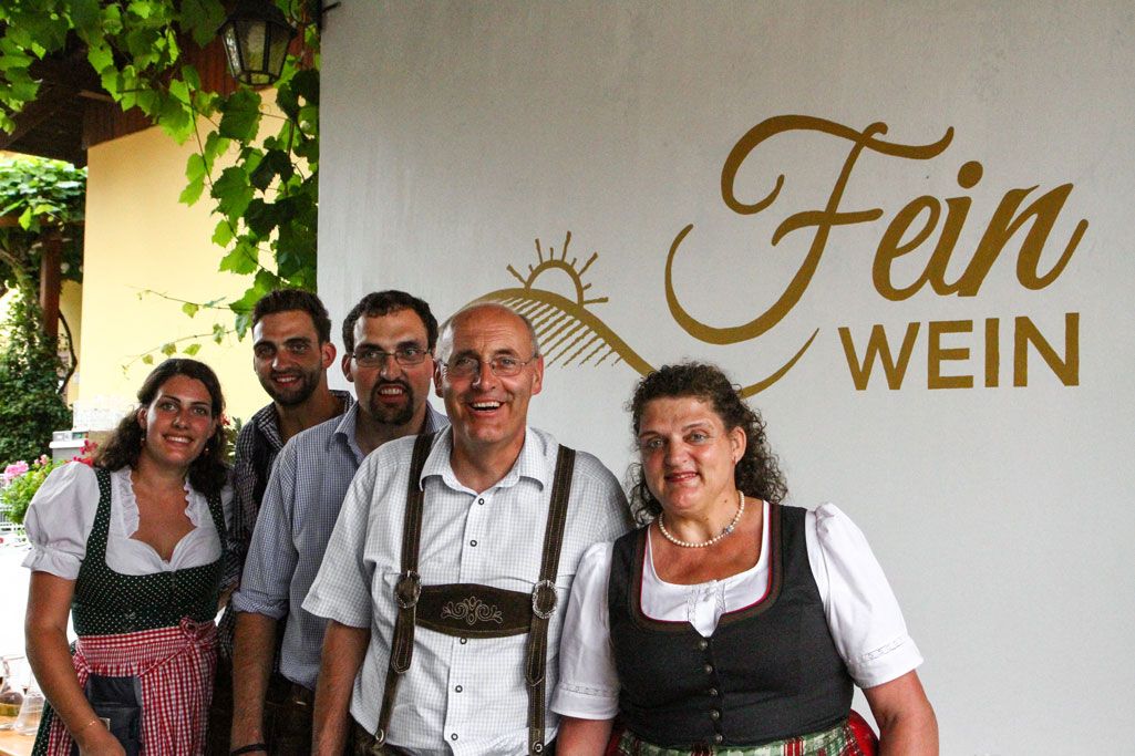 Öt emberből álló csoport hagyományos öltözékben egy "Fein Wein" feliratú tábla előtt.