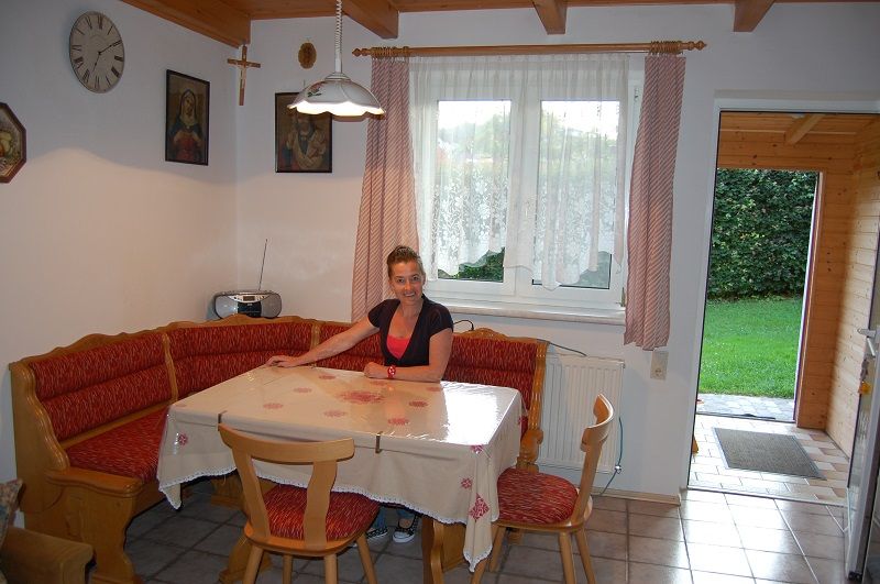 Egy nő ül az étkezőasztalnál egy hangulatos konyhában, sarokpaddal és piros párnákkal.