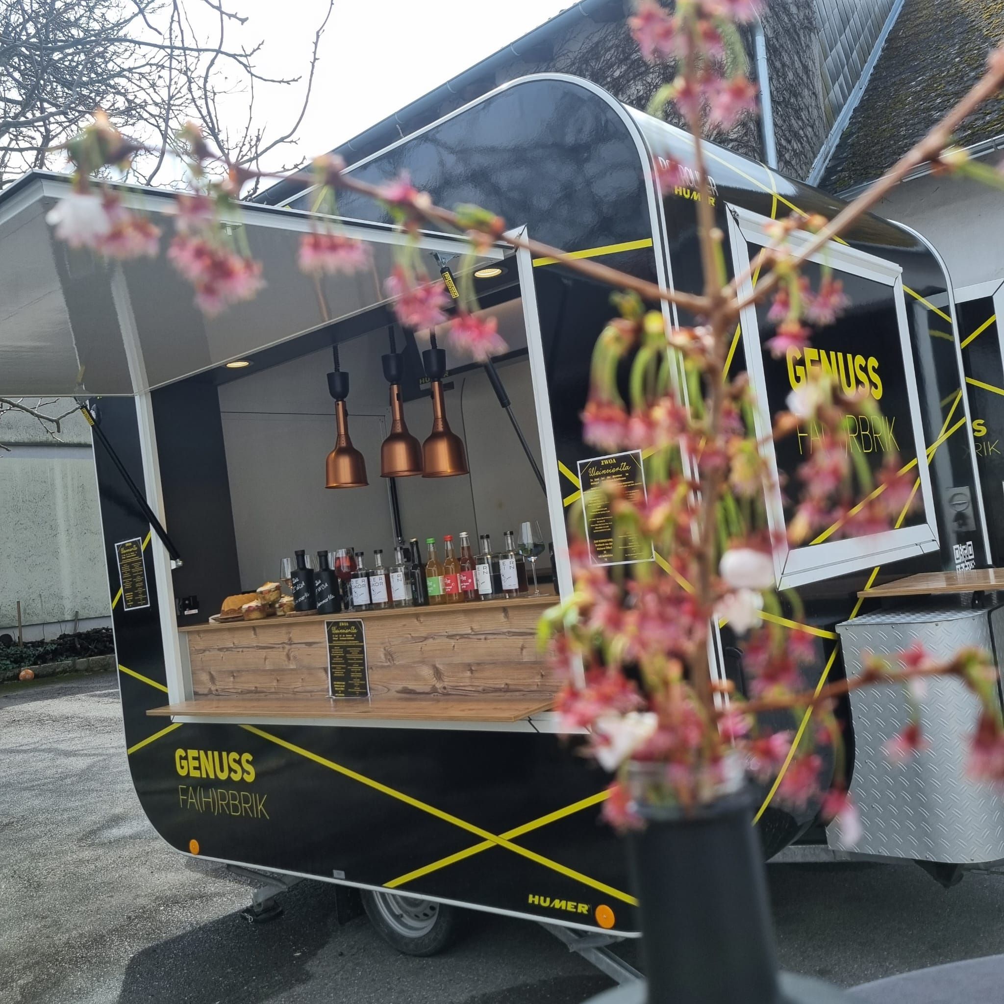 Kint egy fekete ételszállító kocsi parkol "Genuss Fahrbrik" felirattal. Az előtérben elmosódott virágok láthatók.