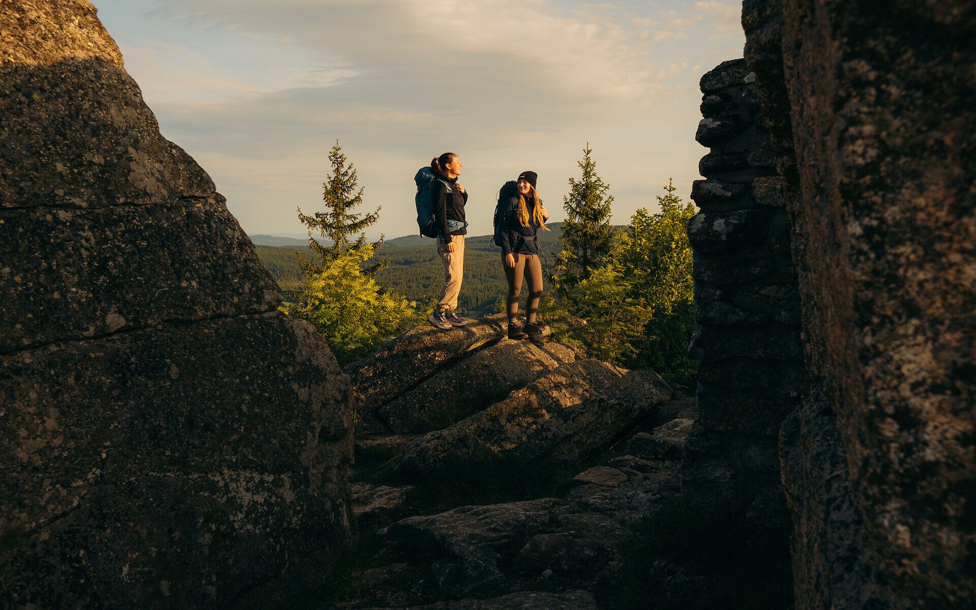 Két túrázó áll az impozáns sziklaalakzatok között, és a Waldviertelben élvezik az erdős táj pazar kilátását az aranyló reggeli fényben.
