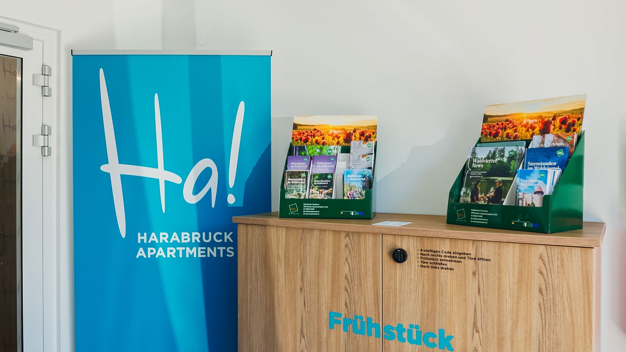 Egy recepciós terület, ahol a Harabruck Apartments kék szalagcímkéje látható, valamint egy prospektustartó áll egy fából készült szekrényen.