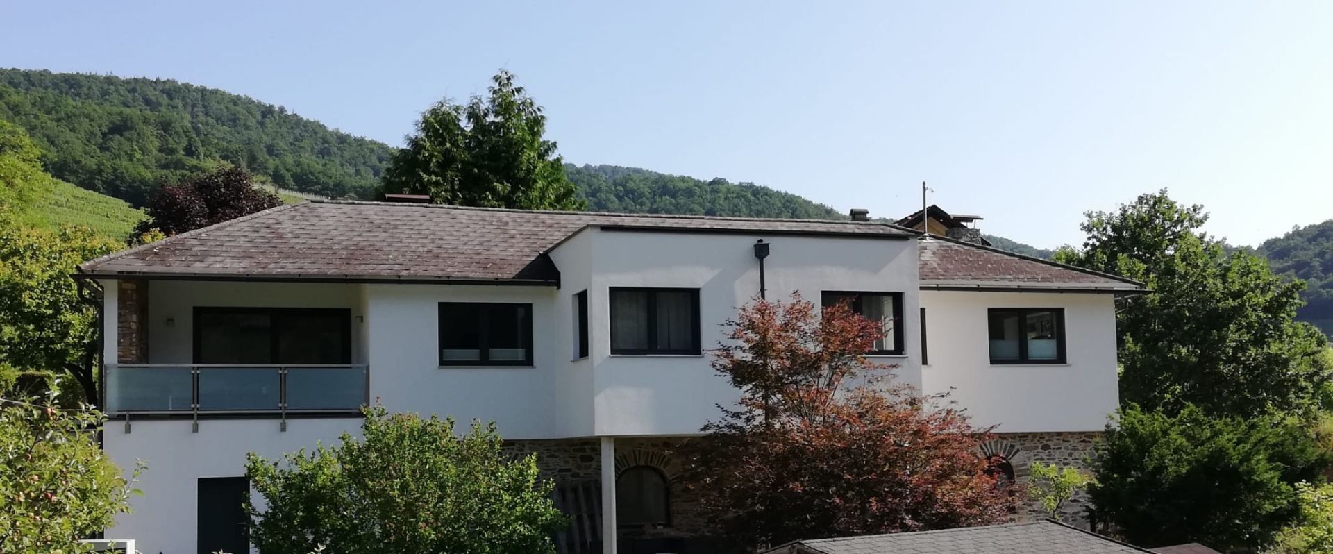 Egy modern fehér nyaraló külső nézete, előtérben fákkal, háttérben erdős dombokkal.