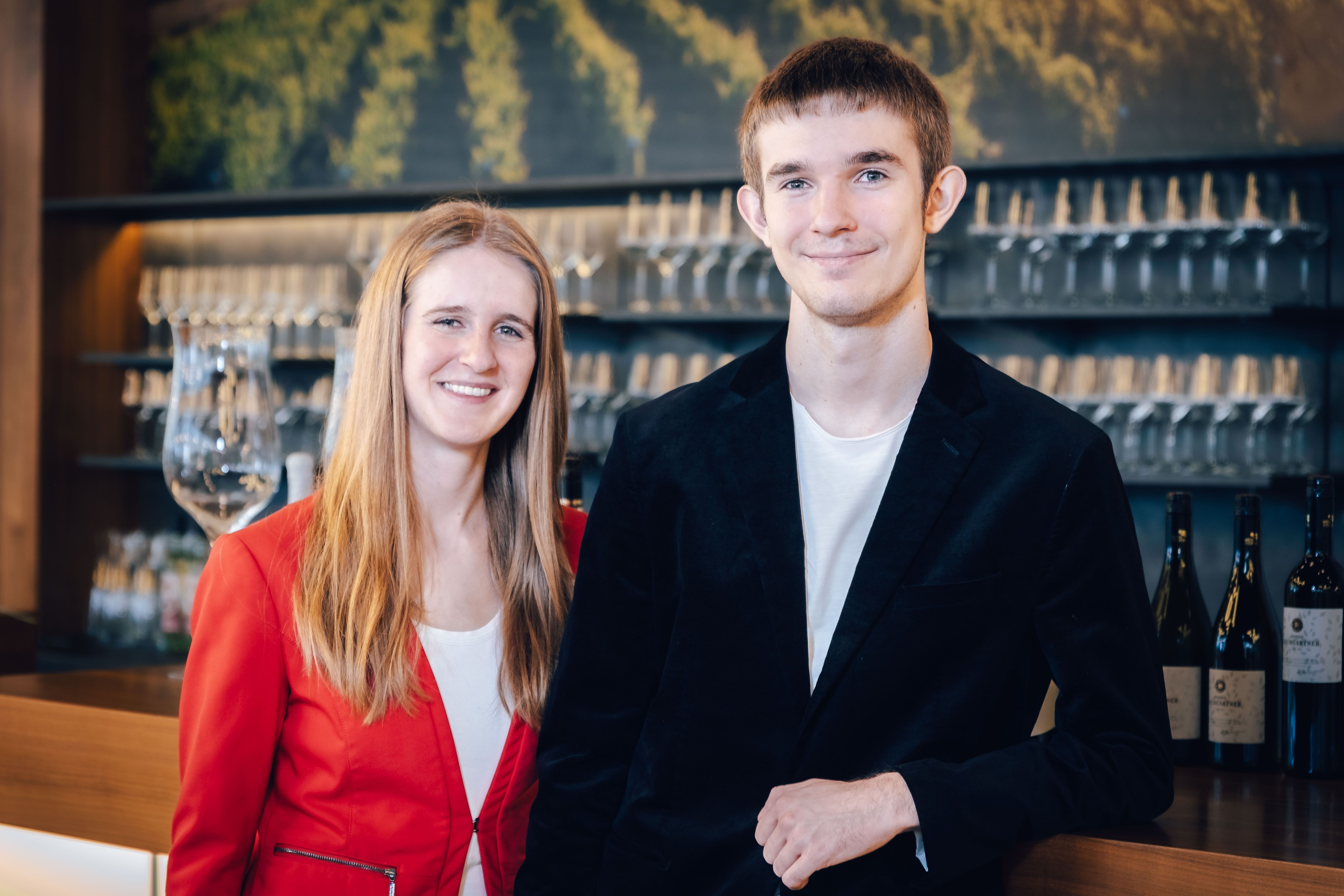 Katharina és Lorenz Baumgartner elegáns ruhában egy borospoharakkal teli polc előtt.