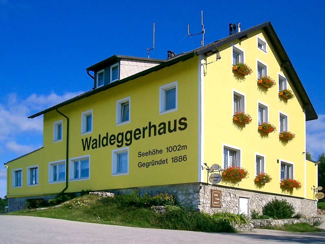 Sárga épület "Waldeggerhaus" felirattal, virágokkal körülvéve.