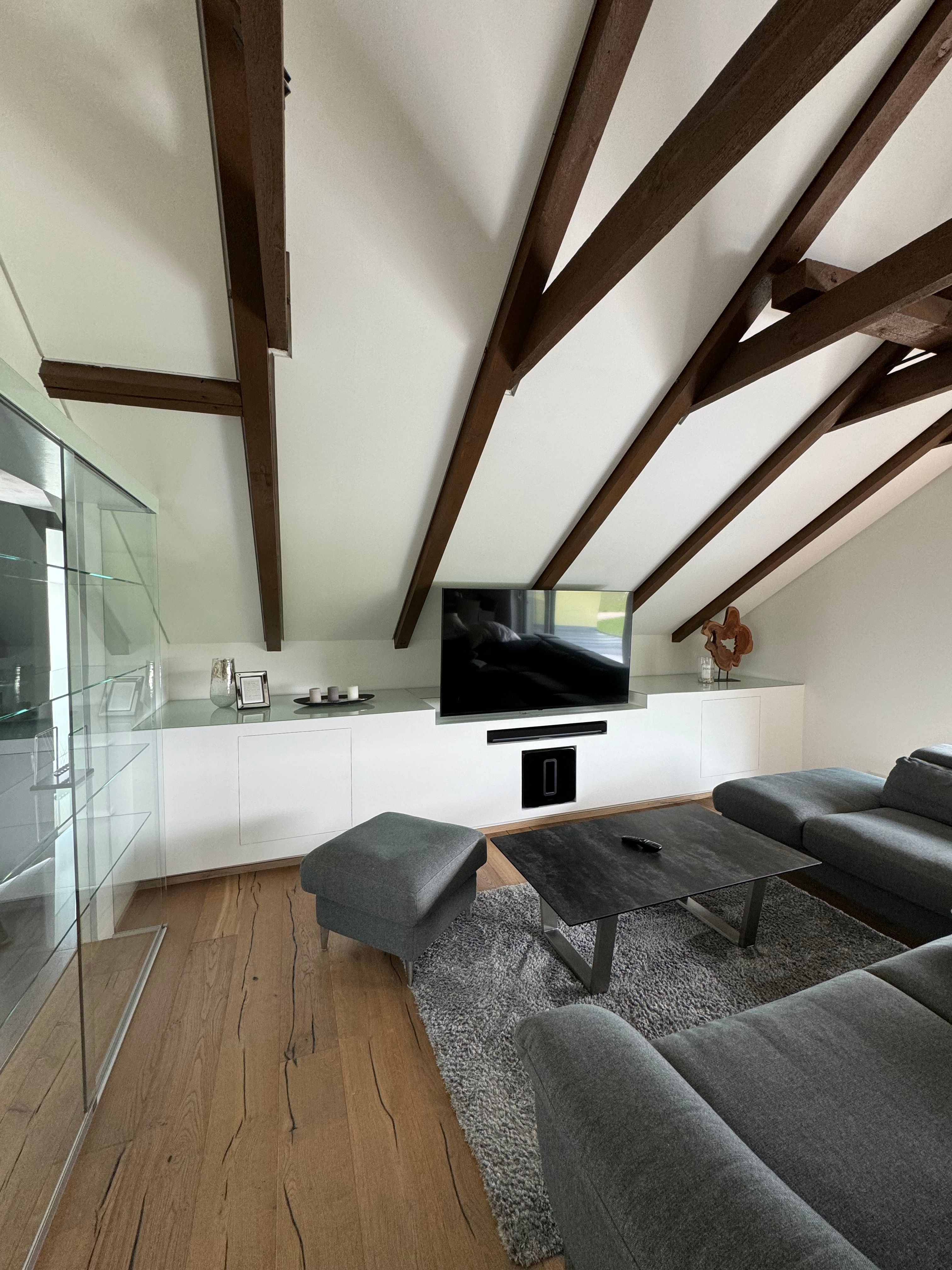 Modern nappali szürke kanapéval, fagerendás mennyezettel és síkképernyős TV-vel.