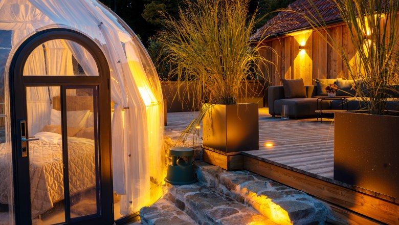 Egy modern faház terasszal és egy átlátszó kupolás sátorral az erdőben alkonyatkor.