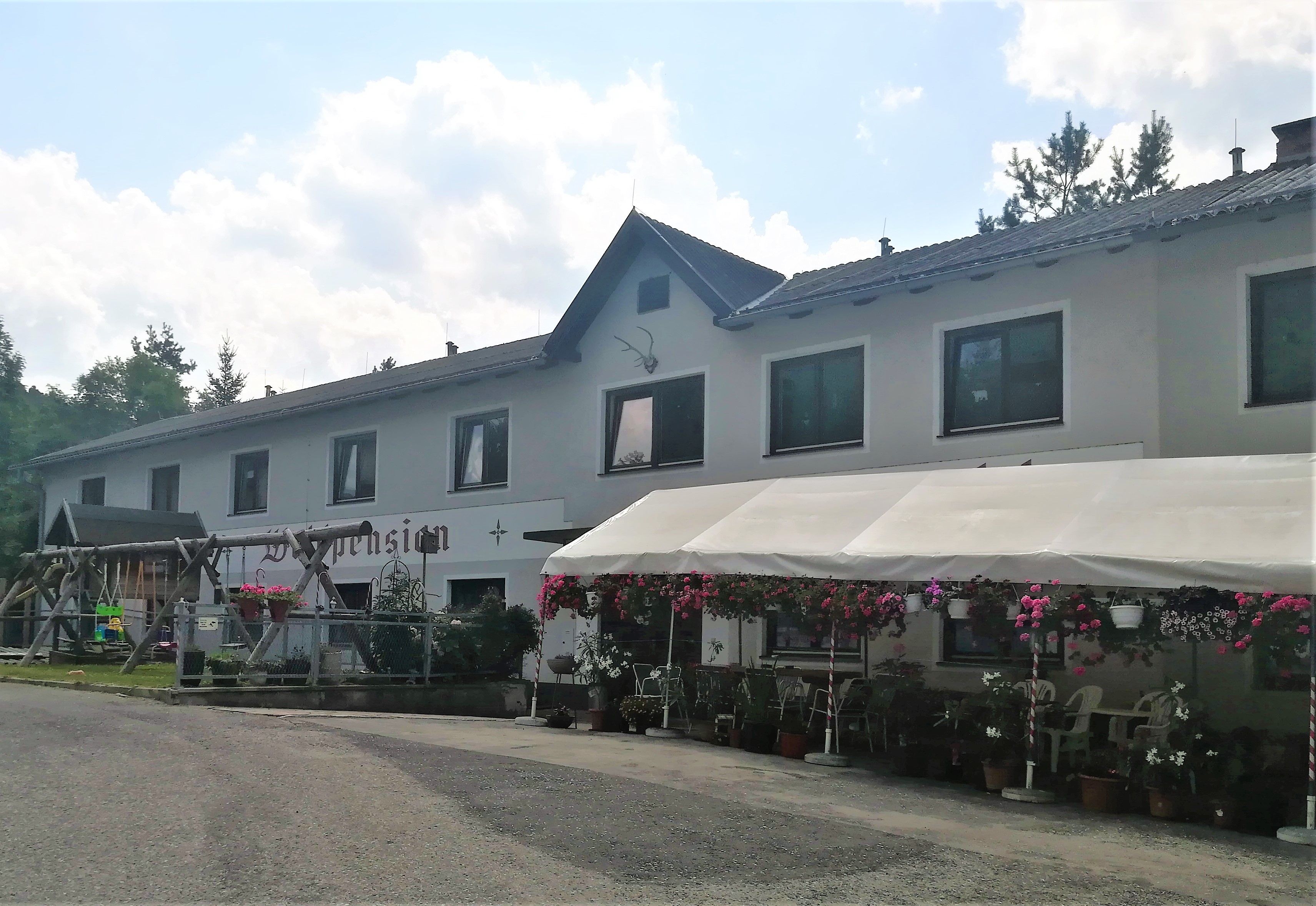 Egy kétszintes, "Waldpension" feliratú épület, körülötte virágokkal és előtetővel.