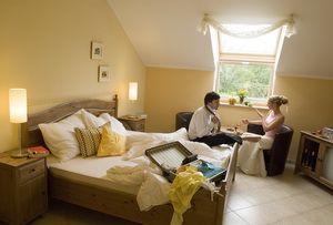 Egy hangulatos szállodai szoba, ahol egy pár ül az asztalnál. Az ágy bevetetlen, és egy bőrönd nyitva hever az ágyon.