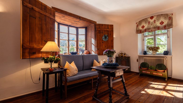 Hangulatos nappali fapadl&oacute;val, kanap&eacute;val, asztallal &eacute;s l&aacute;mp&aacute;val egy nagy, fa redőny&ouml;s ablak előtt.