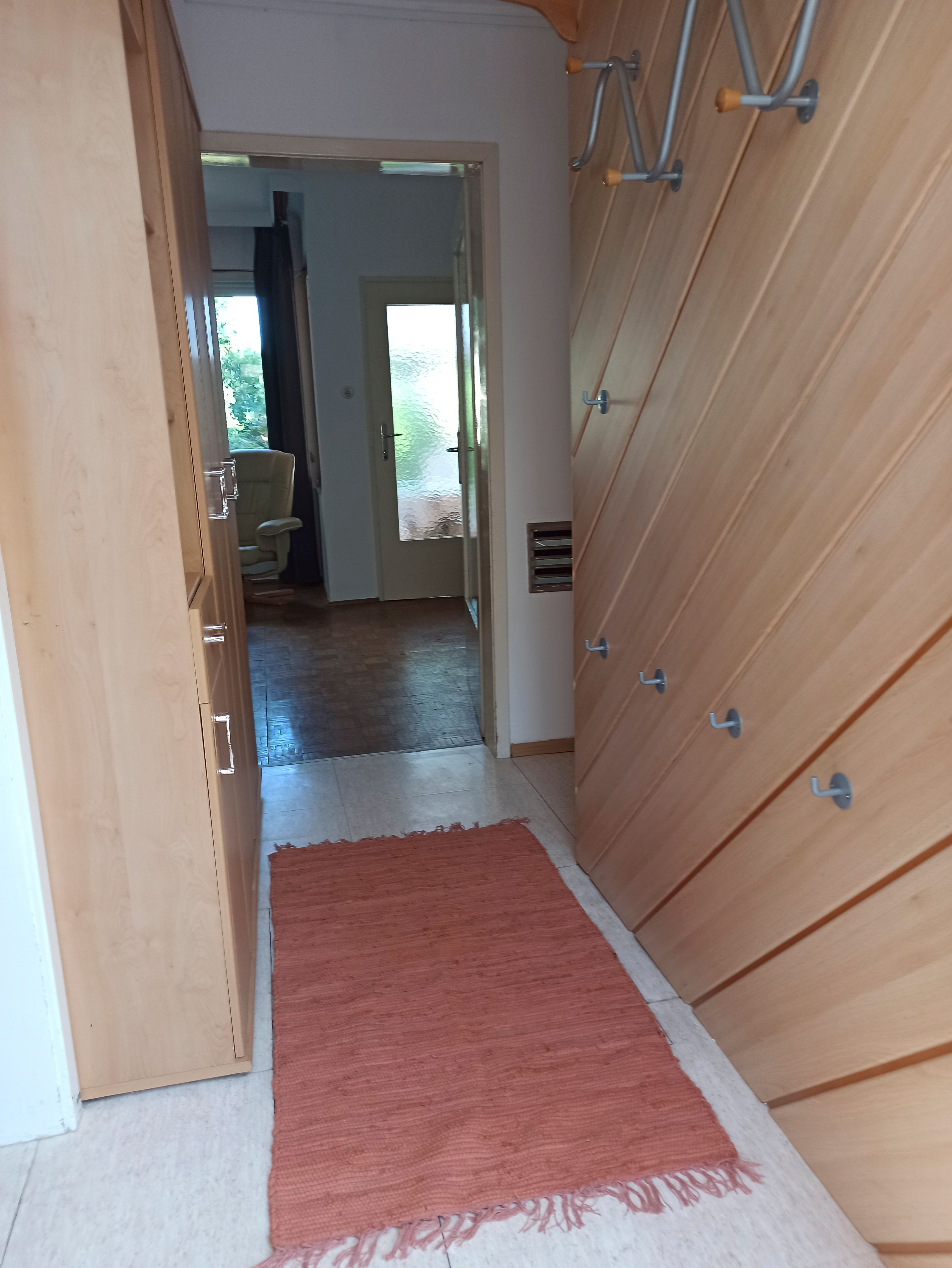 Egy folyosó fából készült falakkal, kampókkal és vörös szőnyeggel. A háttérben egy szoba látható egy karosszékkel és egy üvegajtóval.